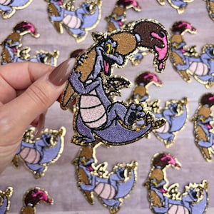 May include: Embroidered patch featuring a purple dragon with a brown paintbrush and pink paint. The dragon has yellow eyes and a gold glitter outline. The patches are on a light wood grain surface.
