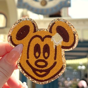 May include: A gold glitter-edged embroidered patch of Mickey Mouse with a brown outline and a yellow face. The patch is held in a hand.