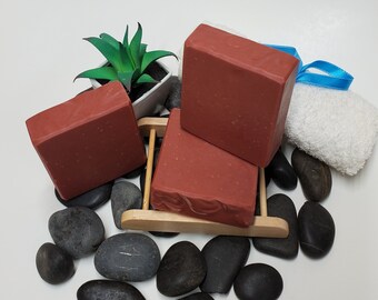 May include: Three red clay soap bars on a wooden stand with black river rocks surrounding the stand. A small potted plant is in the background.