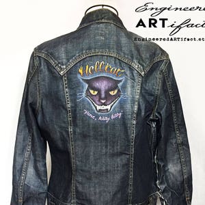 May include: A dark blue denim jacket with a painted design on the back. The design features a black cat with yellow eyes and the words "Hellcat" and "Here, kitty kitty".
