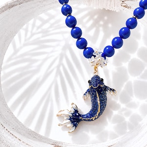 Könnte beinhalten: Halskette mit blauem Koi-Fisch-Anhänger und blauen Perlen. Der Anhänger hat goldene Details und weiße Akzente. Die Halskette besteht aus großen, runden blauen Perlen und kleinen weißen Blumenakzenten.