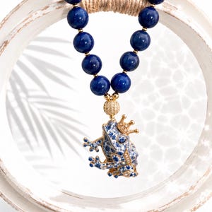 Könnte beinhalten: Eine Halskette mit dunkelblauen, kugelförmigen Perlen und einem goldfarbenen Anhänger in Form eines Frosches mit Krone. Der Froschanhänger ist mit einem blau-weißen Blumenmuster und kleinen klaren Steinen verziert und hängt an einer goldenen Perle.