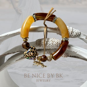 Può includere: Un braccialetto di perline con perline curve ambra e marroni, distanziatori dorati e fasce con strass. Un ciondolo a forma di cuore con stampa leopardata e un ciondolo a forma di tigre dorata pendono dal braccialetto. Il testo "BE NICE BY BK JEWELRY" è visibile.
