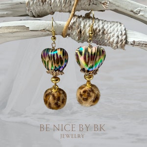 Könnte beinhalten: Ein Paar goldene herzförmige Ohrringe mit Leopardenmuster und einem flauschigen braunen Bommel. Die Ohrringe liegen auf einer weißen Holzoberfläche. BE NICE BY BK JEWELRY