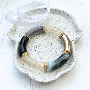 Könnte beinhalten: Ein mehrfarbiges Armband mit schwarzen, grauen, goldenen und durchsichtigen Perlen. Das Armband liegt auf einer weißen Keramikschale mit strukturierter Oberfläche. Die Schale trägt die Aufschrift "Be nice by Bik".