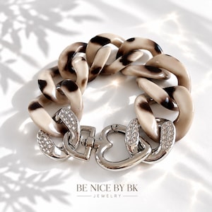 Könnte beinhalten: Ein klobiges Gliederarmband mit beigen und braunen, schildpattfarbenen Acrylgliedern, verbunden mit silberfarbenen Metallgliedern, die mit klaren Strasssteinen besetzt sind, und einem herzförmigen Verschluss. Der Text BE NICE BY BK JEWELRY ist sichtbar.