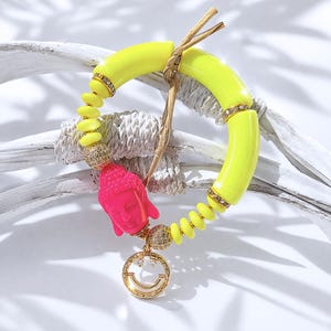 Könnte beinhalten: Ein lebendiges Armband mit neon-gelben, gebogenen Perlen und kleineren gelben Perlen. Ein pinkfarbener Buddha-Kopf-Anhänger und ein goldfarbener Smiley-Anhänger hängen an dem Armband. Das Armband ist mit goldfarbenen Perlen und klaren Kristallen akzentuiert.