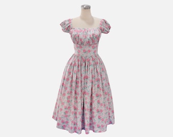 Puede incluir: Un vestido de estilo vintage con estampado floral. El vestido presenta un escote cuadrado, mangas cortas abullonadas y un corpiño ajustado que fluye hacia una falda amplia. La tela es rosa claro con un estampado de rosas y hojas verdes.