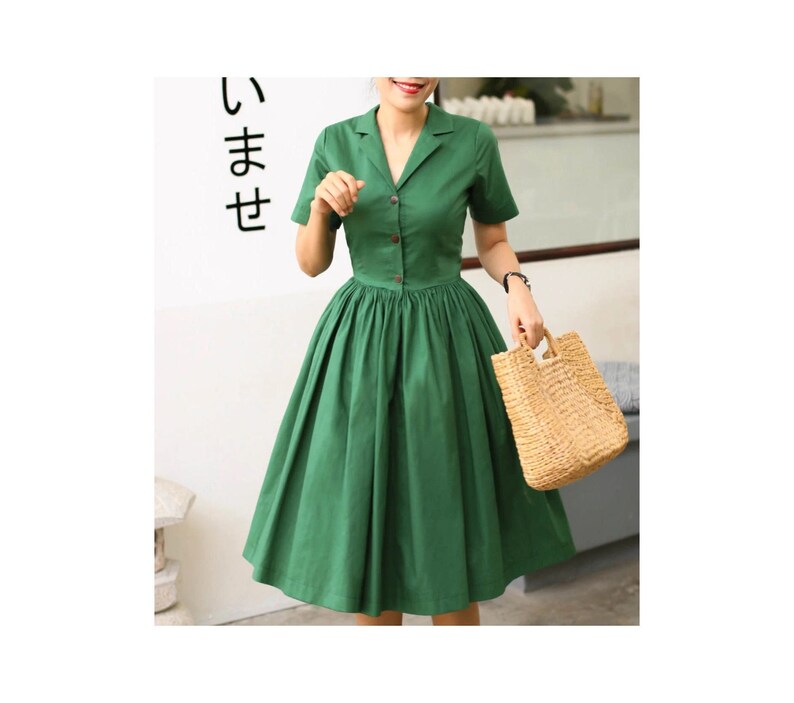 May include: A solid green midi dress featuring a notched lapel collar, short sleeves, and a button-down front closure. The bodice is fitted, transitioning into a full, pleated skirt that falls below the knee. A woven straw handbag is held at the side.