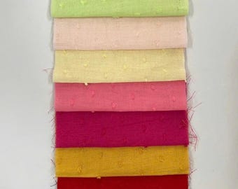 May include: A stack of fabric swatches in various colors, including white, green, pink, yellow, red, and black. Each swatch has a textured, dotted pattern. The fabrics are neatly folded, showcasing the range of hues.