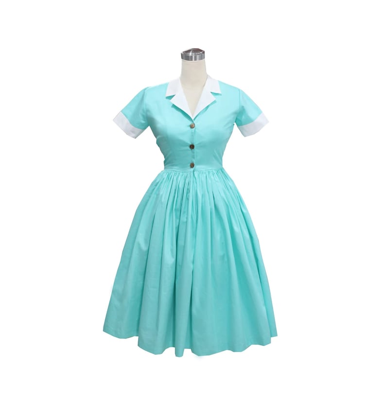 May include: A light turquoise dress featuring a white notched collar and white sleeve cuffs. The dress has short sleeves, a button-down front with three visible brown buttons, and a full, pleated skirt that falls to the knee.