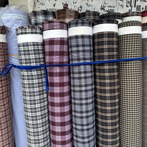 Puede incluir: Una colección de rollos de tela con varios patrones de cuadros y cuadros. Las telas son en tonos de marrón, negro, blanco y burdeos, aseguradas con una cinta azul. Una banda blanca rodea cada rollo.