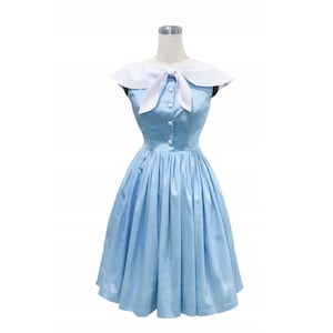 May include: A light blue sleeveless dress featuring a white sailor-style collar with a front tie detail. The bodice includes a vertical row of white buttons down the center, and the skirt is pleated, falling to a knee-length hemline.