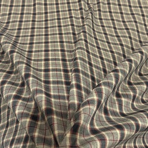 Puede incluir: Primer plano de una tela con estampado de cuadros. La tela presenta un patrón de cuadrícula repetido en azul marino, crema y burdeos sobre un fondo gris. La tela está drapeada, creando pliegues y sombras suaves.