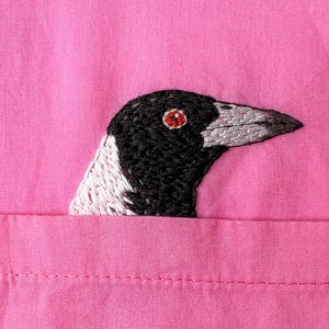 Puede incluir: Primer plano de una tela rosa con un bordado de urraca. La cabeza del ave es negra y blanca con un pico gris y un ojo rojo. La urraca parece estar saliendo de un bolsillo.