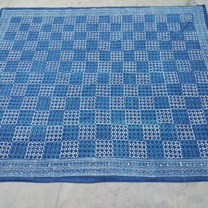 Op de afbeelding: Een grote, indigo blauwe en witte quilt met patroon. De quilt heeft een herhalend dambordpatroon met vierkanten in verschillende tinten blauwe en witte geometrische patronen. De randen zijn afgewerkt met een decoratieve rand.