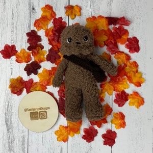 May include: A handmade brown plush toy with a dark brown bandolier, set against a backdrop of autumn-colored leaves. The toy has a smiling face with black eyes. A wooden disc with the text "@SamigurumiDesigns" and Etsy and Instagram logos is in the lower left.