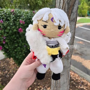 May include: A handmade crochet doll of a character with white hair, wearing a white outfit with a fur-like shawl, and black shoes. The doll has pink cheeks, black eyes, and a purple marking on its forehead. The text "@knotSorsery" is visible.