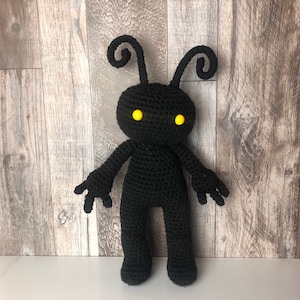 May include: A handmade black crochet doll with yellow eyes and antenna-like features. The doll has a humanoid shape with arms and legs. The background is a light brown wooden surface.
