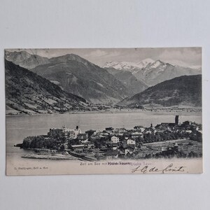 Op de afbeelding: Een vintage zwart-wit ansichtkaart met de stad Zell am See aan een meer met het Hohe Tauern-gebergte op de achtergrond. De tekst luidt Zell am See mit Hohe Tauern, L. Haidinger, Zell a. See en bevat een handgeschreven handtekening.