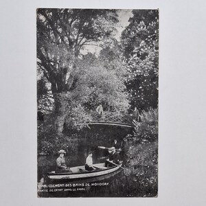 Pode incluir: Postal vintage em preto e branco com uma cena de parque com uma pequena ponte e um barco. O texto na parte inferior diz "ETABLISSEMENT DES BAINS DE MONDORF. PARTIE DE CANOT DANS LE PARC."