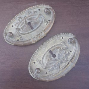 May include: Two oval-shaped brass drawer pulls featuring a central keyhole cutout and a curved handle. Each plate displays a decorative embossed border and a symmetrical relief pattern surrounding the keyhole, with visible screw holes for mounting.