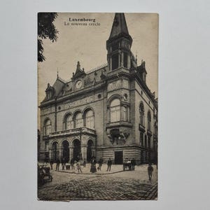 May include: Vintage postcard featuring the "Le nouveau cercle" building in Luxembourg. The sepia-toned image shows a detailed architectural facade with a clock, arched windows, and a tall tower. People and early automobiles are visible in the street.