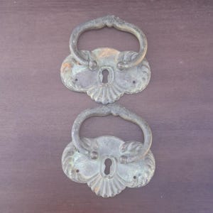 May include: Two matching antique brass drawer pulls with a weathered patina. Each piece features a decorative backplate with a central keyhole and a curved, swinging handle attached at the top. The metal surface shows signs of age and oxidation.
