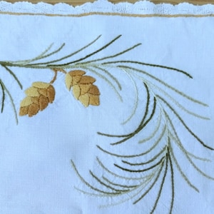 May include: White linen tea towel with embroidered pine needles and pine cones in shades of green and brown.