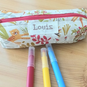 Può includere: Un astuccio per matite in tessuto bianco con chiusura a zip e stampa giungla colorata con tigri, foglie e uccelli. L'astuccio ha un'etichetta con il nome ricamata personalizzata che recita "Louise". In primo piano sono visibili tre pennarelli: rosso, giallo e blu.