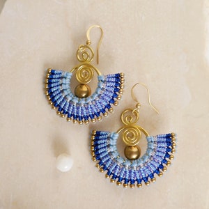 Puede incluir: Un par de pendientes de aro dorados con detalles de macramé azul y morado y cuentas doradas. Los pendientes tienen un diseño en espiral en el centro.