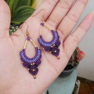 Puede incluir: Pendientes de macramé de color morado y dorado con forma de lágrima. Los pendientes están hechos con una variedad de nudos y cuentas.