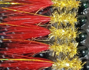 May include: A close-up of six red and gold fishing lures with green beads. The lures have a long, thin, red feather-like material and a shorter, gold, sparkly material.