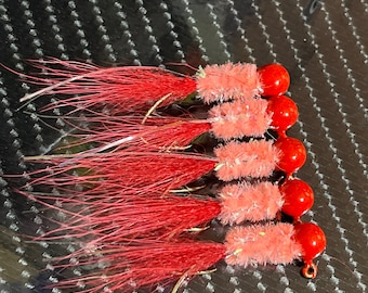 May include: A set of six red and pink fishing lures with a fluffy texture. The lures have a red bead at the end and a hook.