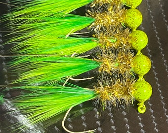 May include: A close-up of six green and gold fishing lures with a fuzzy green and gold body and a round green head.