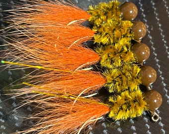May include: Five orange and yellow fishing lures with brown beads. The lures have long, fluffy orange tails.