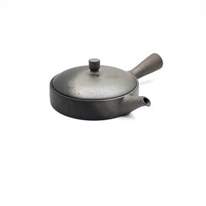 May include: A low-profile, dark gray ceramic teapot with a speckled, matte finish. It features a flat, circular lid with a small cylindrical knob, a short, tapered spout, and a long, slender handle extending from the side of the round body.