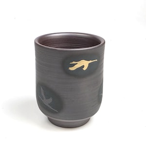 May include: A dark gray ceramic tumbler with a slightly flared base. The tumbler features gold-colored bird silhouettes on a dark green background. The interior is a dark brown color.