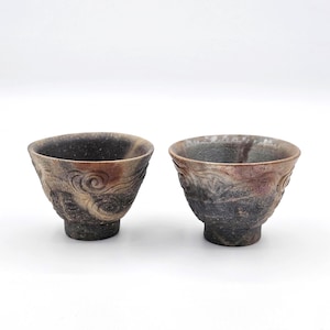 May include: Two handcrafted ceramic bowls with a textured, swirling design. The bowls have a dark, speckled exterior with hints of brown and red. The bowls are small and have a footed base.