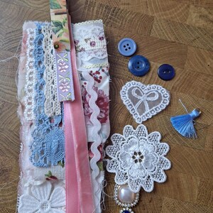 Può includere: Una collezione di materiali per l'artigianato in tonalità pastello. L'immagine presenta pizzi, nastri, bottoni e abbellimenti. Una molletta con motivo floreale tiene insieme dei campioni di tessuto. Ci sono bottoni blu, un pizzo a forma di cuore e un nappa blu.