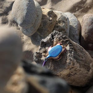 May include: A blue and purple wooden turtle pendant with a beaded cord. The pendant is sitting on a rock.