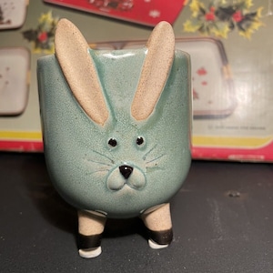 May include: A ceramic planter shaped like a rabbit with long ears, a light blue glaze, and black eyes. The planter has white feet with black accents.