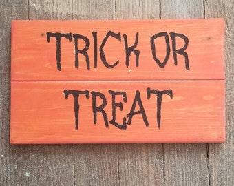 Könnte beinhalten: Oranges Holzschild mit den Worten "TRICK OR TREAT" in schwarzer Schrift. Das Schild besteht aus zwei horizontalen Holzbrettern und eignet sich für Halloween-Dekorationen.