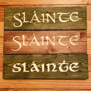 Peut inclure: Trois panneaux en bois avec le mot irlandais "Sláinte" écrit en peinture blanche. Les panneaux sont empilés les uns sur les autres, le panneau du haut étant vert, le panneau du milieu étant brun et le panneau du bas étant vert.