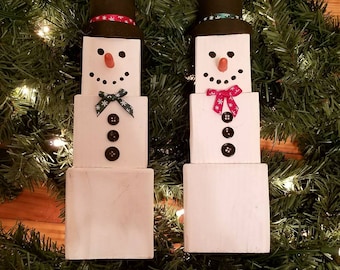 Puede incluir: Dos decoraciones de muñeco de nieve de madera, cada una hecha de cuatro bloques apilados. El bloque superior es un sombrero negro con un patrón de copo de nieve blanco. El segundo bloque es una cara blanca con ojos negros, un lazo rojo o verde y una nariz marrón. El tercero tiene tres botones negros. El bloque inferior es blanco. Los muñecos de nieve están sentados en una guirnalda verde.