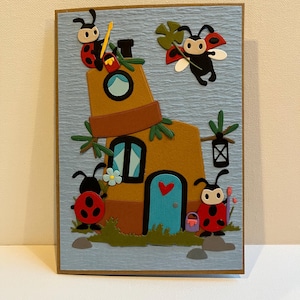 May include: A whimsical handmade card featuring a brown house with a blue door and windows, adorned with ladybugs. The card has a blue textured background and a brown border. Ladybugs are depicted in various poses, adding a playful touch.
