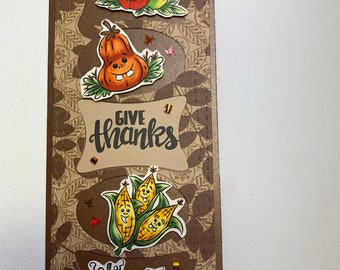 May include: A brown card with a fall theme. The card has a brown background with a leaf pattern. The card has a variety of fall fruits and vegetables with faces on them. The card says "Give Thanks" in a brown cursive font.