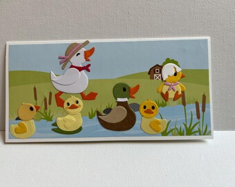 May include: A card with a white duck wearing a hat, a brown duck, a yellow duck, and two yellow chicks. The ducks and chicks are all in a pond with green reeds.