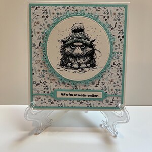 May include: Greeting card featuring a grumpy cat wearing a winter hat, with the text "Not a fan of sweater weather." The cat illustration is in a circular frame, set against a floral patterned background with a teal border. The card is displayed on a clear plastic stand.