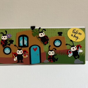 May include: A whimsical card featuring a brown house with a blue door and round window, decorated with ladybirds. The phrase "Cute as a bug" is written on a yellow oval. The background is green.